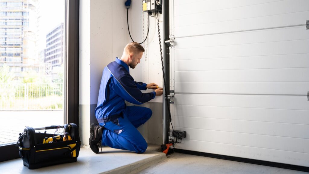 A Technician in a Blue Jumpsuit Kneels Beside a Garage Door Track He is Installing.