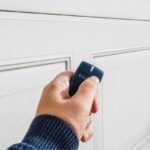 A hand holding an older style garage door opener in front of a garage door.