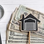 A stack of money next to a magnifying glass and a small wooden house with a garage.