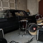 A garage interior with a car and band equipment, including a drum set and amplifiers.