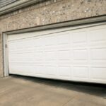 A crooked white garage door, slightly open, attached to a beige brick house.