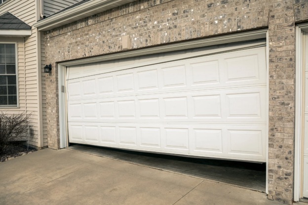 A crooked white garage door, slightly open, attached to a beige brick house.
