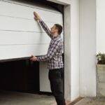 Man in a flannel shirt checking a white, halfway open garage door outside a house.