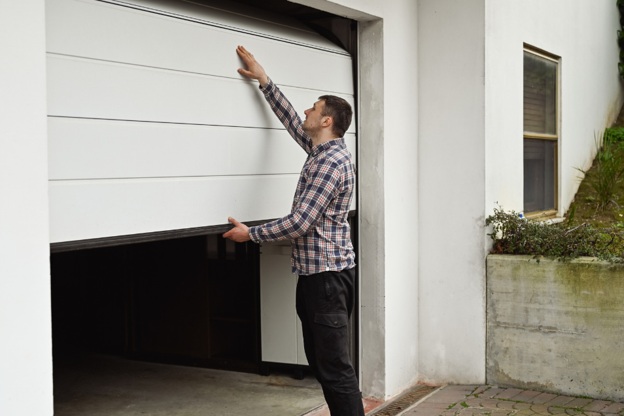 Man in a flannel shirt checking a white, halfway open garage door outside a house.