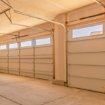 Spacious two-car garage interior with closed sectional garage doors, overhead door tracks, drywall-finished walls and ceiling, and a clean concrete floor illuminated by natural light from upper door windows.
