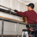 Technician in a dark red shirt uses tools on a black torsion spring above a textured white garage door in a bright garage.