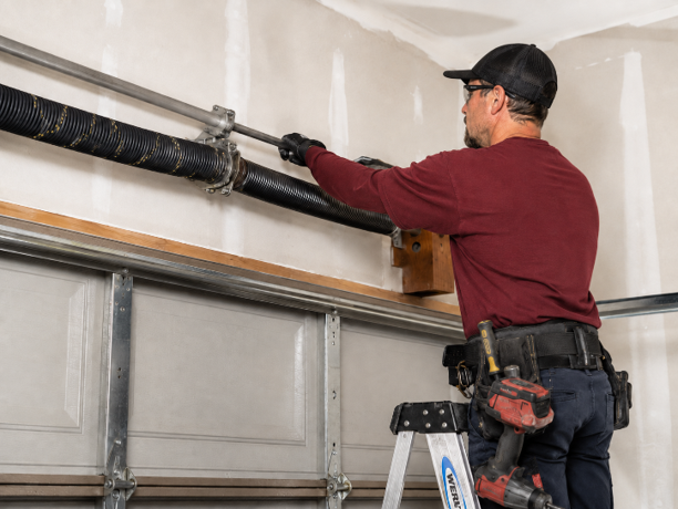 Technician in a dark red shirt uses tools on a black torsion spring above a textured white garage door in a bright garage.