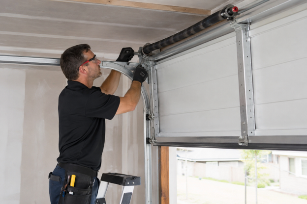 Technician in a black shirt and gloves repairs silver garage door tracks and springs beside textured white metal garage door panels.